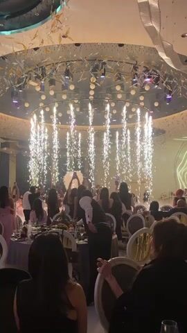 A celebratory wedding scene unfolds indoors, capturing the first dance of the newly married couple. The venue is an opulent banquet hall with guests seated around tables, many recording the event with their phones. Suspended above the dance floor is a large, circular chandelier emitting bright sparks, creating a dazzling effect as they descend. The bride and groom are the focal point, visible amidst the gathering of people, sharing their first dance. The room's decor includes intricate overhead decorations, adding to the grandeur of the occasion. A waiter is seen tending to a nearby table.