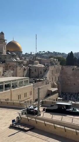 The video shows an aerial view of the Western Wall in Jerusalem. In the foreground, there is a large paved plaza. In the middle ground, there is a raised platform with a black surface and a white object that appears to be a table or altar. There are several people gathered at the Western Wall, which is a large ancient stone wall. Some people are wearing white clothing. In the background, there are buildings and trees. The sky is clear and blue.