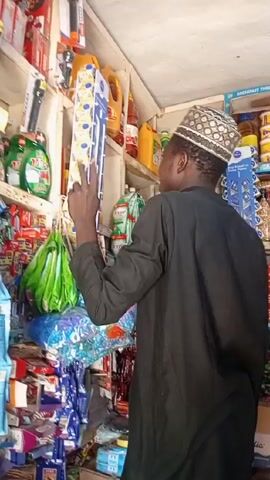 A man in a black shirt and a patterned cap is standing in a small, densely packed store. He's reaching up to a shelf to grab a package of small, round containers, possibly yogurt or a similar snack. The shelves around him are crammed with various items, including flashlights, detergents, and other packaged foods. The lighting in the store is bright, and the scene appears to be indoors.