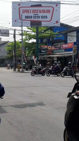 The video captures a street scene with a billboard in the background that reads "SPACE DISEWAKAN" and a phone number. Several motorcycles are visible on a paved road, some of which are in motion. One motorcycle rider on the left is turning left, another rider is going straight ahead, and a third rider is also moving forward. The environment appears to be an intersection or a busy street with buildings and trees in the background. The lighting suggests it is daytime.