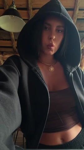 A young woman with long dark hair, wearing a black zip-up hoodie over a brown crop top and black sweatpants, stands indoors. She adjusts her hoodie and necklace, touching her face and hair. The environment appears to be a rustic indoor space with wooden beams and exposed ceiling structures, two industrial-style hanging lamps, and glimpses of outdoor greenery through a large window or opening. Chairs and tables are partially visible in the background. The woman makes various gestures and poses, looking towards the camera.
