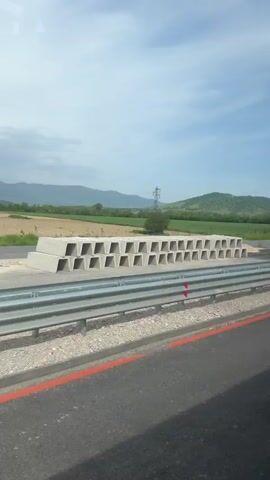 The video shows a green Scania truck parked next to a stack of concrete barriers along a roadside. The truck has a Hulk figure visible on the windshield. In the background, there's a field, trees, and a mountain range. The sky is cloudy. The truck's license plate is visible. The video appears to be taken from inside a vehicle.