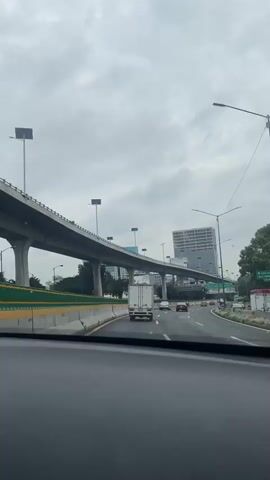 The video shows a view from inside a vehicle driving on a multi-lane highway. The highway has an elevated section to the left, supported by concrete pillars. The sky is overcast with gray clouds. Several vehicles are visible on the highway, including a white truck, passenger cars, and a black car. There are streetlights and some trees and buildings in the background. The road appears to be wet, suggesting it may have rained recently.