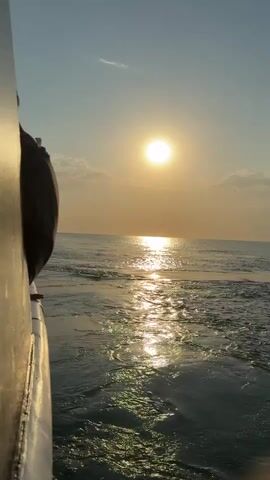 The video shows a person sitting on the side of a boat looking out at the ocean during sunset. The sun is a bright orb in the sky, casting a reflection on the water. The boat is moving, creating a wake behind it. The sky is clear with a few wispy clouds. The person is mostly obscured by the side of the boat, but appears to be wearing dark clothing. The overall scene is peaceful and serene.