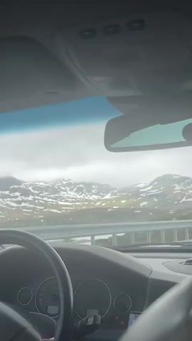 The video shows the interior of a car, looking out through the windshield. The driver's hands are visible on the steering wheel. Through the windshield, a mountainous landscape with patches of snow can be seen under a cloudy sky. A metal guardrail runs along the side of the road. The dashboard of the car is also visible, with its gauges and controls. There is a phone mounted on the dashboard with a navigation app showing a map and a speed limit icon indicating 80. The overall mood appears to be one of travel and exploration. The weather appears to be overcast or rainy, with water droplets visible on the windshield. The time of day is unclear due to the overcast conditions, but it is likely daytime. There is a Volvo logo visible on the steering wheel.