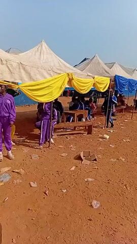 The video shows an outdoor event with multiple tents and people. The ground is red soil with scattered debris. There are several tents with white tops and colored fabric draped underneath, including yellow and blue. People in purple tracksuits are visible, standing and sitting around wooden tables under the tents. A child in jeans and a white tank top walks through the scene. There are adults wearing casual clothing, and at one point, a man in a black suit approaches people under a yellow-draped tent. The atmosphere appears busy and festive.