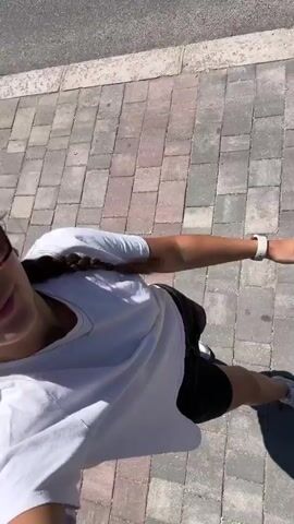 The video begins with a close-up of a young woman from the chest up. She is outdoors, with cobblestone paving visible beneath her and a sidewalk to her left. She has her hair in a braid and is wearing a white t-shirt and black shorts. As the video progresses, she turns her head towards the camera, smiling and wearing sunglasses. The background shows a street with buildings and trees. She then puckers her lips towards the camera.