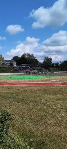 The video shows an outdoor track and field stadium on a sunny day with a blue sky and white clouds. In the foreground, there is a chain-link fence. Beyond the fence is a grassy area and then a red running track. Further in the background, there is a large green tarp spread out on the field, possibly for an event. There are also bleachers and some people in the distance. The scene appears to be set up for a sporting event or an outdoor gathering.