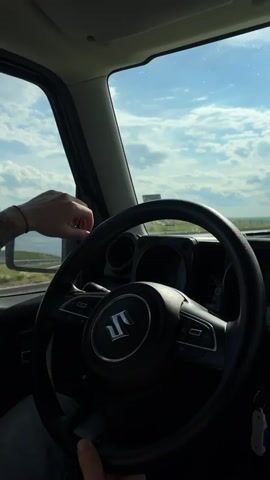 The video depicts the interior of a car from the driver's perspective. The main focus is on the steering wheel, which prominently displays the Suzuki logo. A hand is visible resting on the steering wheel. Outside the vehicle, a highway stretches into the distance under a partly cloudy sky. The car appears to be in motion.