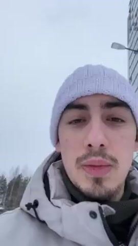 Here is a description of the video.

A man wearing a white beanie and a gray jacket stands in the snow, looking at the camera. He has brown hair and a goatee. The background shows a snowy landscape with trees. The camera then shifts to show two children sledding down a snowy hill. One child is in front and the other is holding on behind. The camera then returns to the man's face.  He appears to be looking on at the children sledding.