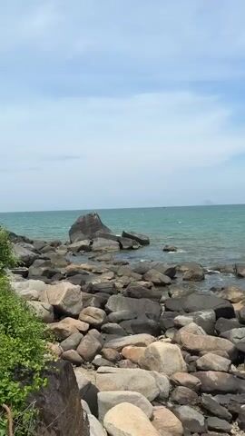The video shows a rocky coastline with the ocean in the background. The sky is partly cloudy. There are several boats visible on the water.