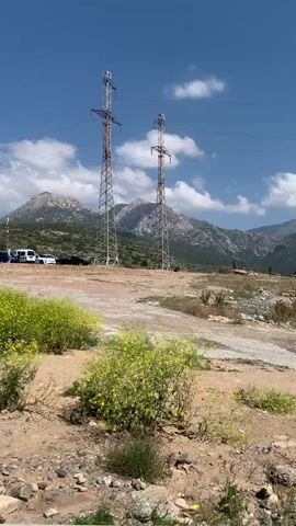 The video captures an outdoor scene with a bright blue sky and scattered white clouds. In the foreground, there are patches of dry, reddish-brown soil with sparse, yellow flowering bushes and green shrubs. Two tall, metal electrical transmission towers stand prominently in the middle ground. Beyond the towers, a winding dirt path or road is visible. In the distance, a range of mountainous terrain unfolds. The mountains are covered in green vegetation on their lower slopes, transitioning to rocky, barren peaks, some of which are capped with snow. Several vehicles, including what appear to be white SUVs and dark-colored cars, are parked in the upper left portion of the frame, near the base of the mountains. A few figures, appearing as small silhouettes, are scattered across the landscape in the middle and far distances. The overall environment suggests a rural or remote mountainous area on a clear, sunny day.