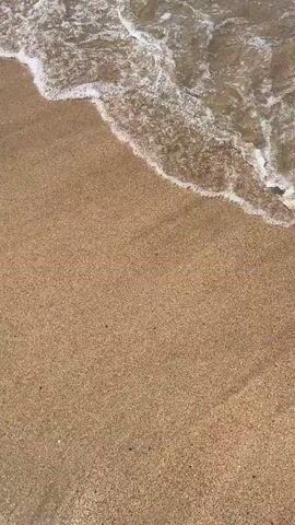 A video shot from a first-person perspective looking down at a sandy beach and the shallow water approaching it. The water is murky brown and somewhat foamy as it washes over the sand. The sand is a light brown color with small grains.