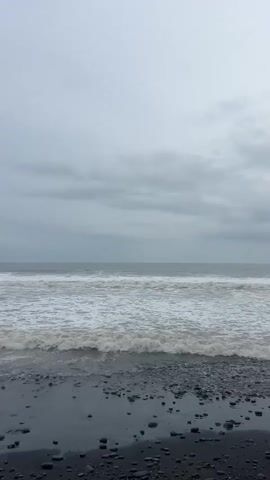 A vertical, low-angle shot shows waves rolling onto a dark, pebbly beach under a heavily overcast sky. The sea is a deep gray with whitecaps and foamy surf, and the horizon is a hazy line separating the sea from the clouds. The beach in the foreground is composed of dark, smooth pebbles and wet sand. No people or animals are visible. The overall mood is somber and desolate.