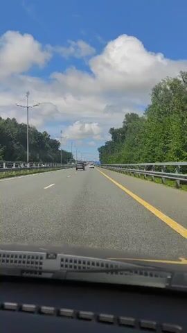 The video shows a forward-facing view from inside a vehicle traveling on a highway. The highway has multiple lanes, separated by white lines, and a prominent yellow line marking the right edge of the lane. On either side of the highway are dense green trees, and a metal guardrail runs along the right side. The sky is blue with fluffy white clouds. Several cars are visible ahead on the highway, moving in the same direction. The lower part of the windshield and the windshield wipers are visible in the foreground. The overall impression is of a sunny day drive on a well-maintained highway surrounded by nature.