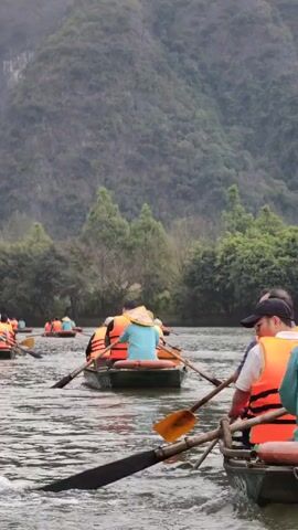 The video shows several small boats on a river, each carrying a group of people.  The people are wearing orange life vests and are using oars to propel the boats through the water.  Some are wearing hats.  The background consists of lush green trees and a large mountain covered with foliage under a cloudy sky. The river appears calm.