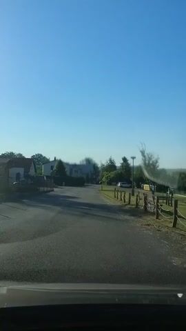 The video is a first-person perspective, likely from inside a vehicle, looking out at a quiet, rural road. The sun is shining brightly in a clear blue sky, casting long shadows across the asphalt road. On the left side of the road, there are several houses with roofs visible, some with trees and other greenery around them. There is a parked car visible in front of one of the houses. On the right side of the road, there is a fence made of wooden posts and rope, with some yellow bins or containers behind it. Further down the road on the right, there are more trees and what appears to be a utility pole. The overall scene suggests a peaceful, everyday setting in a residential or rural area during daylight hours. There are no people or discernible actions taking place in the video.