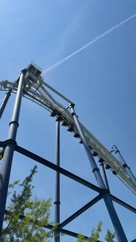 A roller coaster train with several passengers is ascending a steep incline on a bright, clear day. The train is positioned on a white track supported by dark blue, metal structures. The sky is a vibrant blue with a faint white contrail from an airplane. Green foliage from trees is visible at the bottom of the frame.