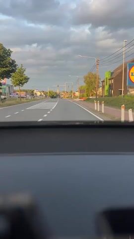 A first-person perspective video shows a road with a crosswalk and white dashed lines. There are houses and trees on either side of the road, with power lines and streetlights overhead. The sky is cloudy. There is a traffic light in the distance that is red. The video is taken from inside a vehicle.