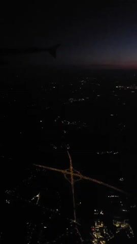 This media file captures an aerial view of a city at night, likely from an airplane. The primary subject is the sprawling urban landscape illuminated by numerous lights, highlighting roads, buildings, and the distinct cloverleaf shape of a highway interchange. The time of day is clearly nighttime, with the sky exhibiting a deep indigo hue transitioning to a faint reddish glow on the horizon, suggesting either dusk or dawn. The weather appears clear, as the lights are sharp and distinct without any atmospheric diffusion.

The scene is set high above the ground, providing a panoramic perspective. The presence of an airplane wingtip in the upper left corner strongly indicates the viewpoint. The location is identified as Witobel, Poland. The overall mood evoked is one of peaceful observation and wonder, seeing the city transformed by artificial light in the darkness. There is no visible text in the media file.