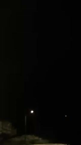 The video shows a dark, outdoor scene at night. A single street light is visible on the left side of the frame, providing a small amount of illumination to the immediate area. In the far distance, on the right side, there is a small, indistinct point of light. Bushes or other foliage are visible along the bottom edge of the frame. The sky is obscured by darkness.