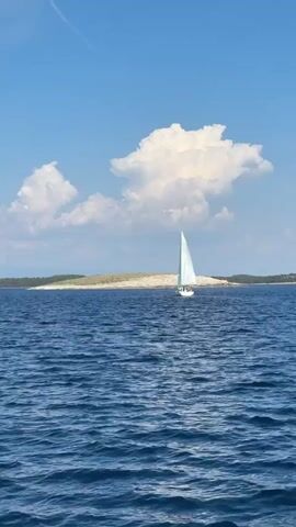 A solitary white sailboat with a full sail glides across a deep blue, rippling ocean under a bright, partly cloudy sky. The boat is positioned off-center to the right, moving towards the left of the frame. In the distance, a sparsely vegetated island with a sandy shoreline and low hills stretches across the horizon. Further left, a more lush, green island with more substantial hills is visible. The overall scene evokes a sense of peacefulness and a serene summer day. The time of day appears to be midday, given the bright sunlight casting clear shadows. The weather is fair with scattered white cumulus clouds against a vivid blue sky, suggesting pleasant sailing conditions. The location is identified as Ćunski, Croatia, which is consistent with the visual elements of islands and the Mediterranean Sea. There is no visible text in the media file.