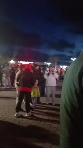 The video depicts a crowd of people gathered in an outdoor area at night. The sky is dark blue, with some clouds visible. There are buildings in the background and streetlights providing illumination. People are wearing various types of outerwear, and some are holding phones. The ground appears to be paved. The video is slightly shaky and out of focus at times. It is difficult to discern specific actions or interactions due to the distance and lighting.