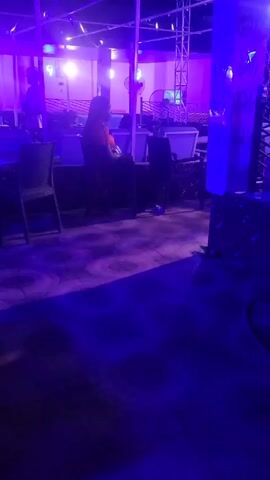 The video shows a room lit with blue and purple light. There are tables and chairs visible, and at least two people in the background. One person is sitting, and the other person is standing behind the table. There is a mist on the floor. The scene appears to be indoors, possibly a nightclub or lounge.