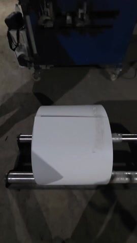 The video shows a machine processing a roll of white material in a warehouse setting. The machine, primarily blue, has a control panel with a screen and an emergency stop button. It also has what appears to be a pump on the left side. The material roll is mounted on a system of rollers and appears to be fed into the machine. Stacks of wrapped material are visible in the background, along with other machinery. The warehouse is dimly lit, but the machine and material are clearly visible. There are no people visible in the video.