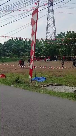 The video shows an outdoor scene of children playing soccer on a field. The field is dirt and grass, with a makeshift soccer goal visible in the background. Several children are actively engaged in the game, kicking and chasing a soccer ball. Spectators, likely adults and other children, are seated or standing around the perimeter of the field, watching the game. The area is decorated with red and white flags, which appear to be strung across the field and on poles. There is a large electrical tower in the background, and trees and vegetation surround the playing area. In the foreground, a paved road is visible, with a patch of grass and dirt between the road and the field. A large red and white patterned flag on a pole stands prominently in the foreground, partially obscuring the view of the game. A blue tarp is also visible on the ground near the flag. The overall impression is a community event, possibly a local celebration or tournament.
