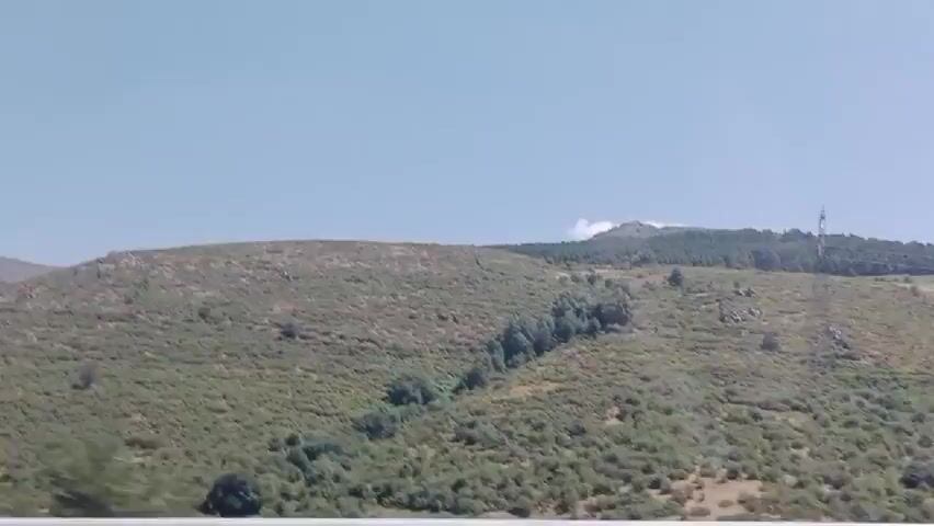 A landscape view from a moving vehicle shows a clear sky over rolling hills covered in green brush and scattered trees. The foreground has a blurry white barrier. A blue road sign with two white arrows pointing upwards is visible on the right. A red and white circular speed limit sign showing "70" is also present on the right. The landscape appears to be mountainous.