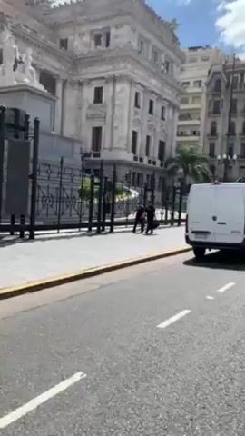 The video shows a street scene in what appears to be a city. A white van is initially in view, parked on the right side of the street with cars passing it. A large, ornate building with a wrought-iron fence is visible on the left side of the street, with a couple of people standing on the sidewalk near the fence. The camera follows the cars as they drive down the street. The buildings lining the street are tall and feature architectural details. The sky is blue with some clouds.