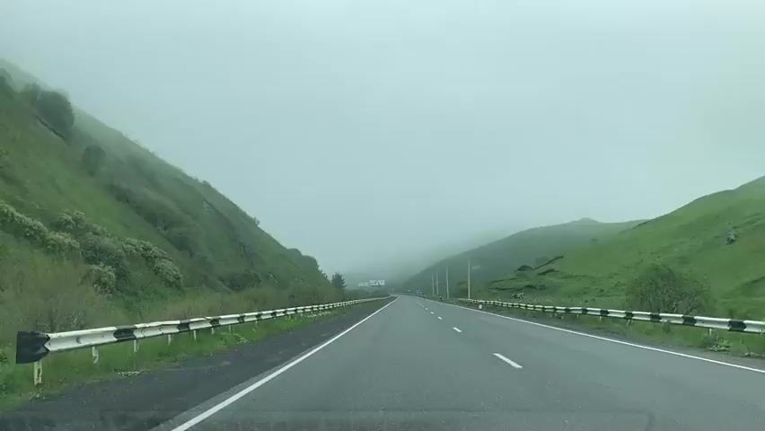 A video shows a forward-facing view from a moving vehicle on a highway. The highway is an asphalt road with a white dividing line and guardrails on either side. The surrounding environment is characterized by lush green, sloping hills on both sides of the road. A thick, white fog or mist obscures the distant landscape, creating a soft, hazy atmosphere. The sky is overcast and uniformly gray. Small trees and bushes are visible along the base of the hills and near the guardrails. The road appears to be in a mountainous or hilly region.