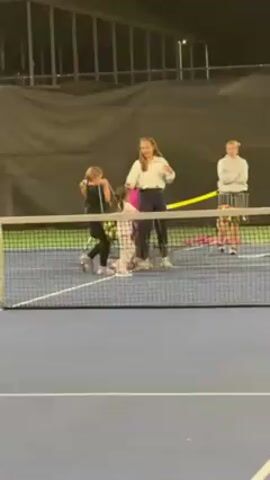 The video shows a group of four young children and two women gathered on a blue indoor tennis court. A tennis net divides the frame horizontally. One woman, with long brown hair and wearing a white sweater, appears to be instructing the children. Another woman, with blond hair, stands with her arms crossed, observing. A black chair with a bag on it is visible near the leftmost child, who is wearing a pink jacket and plaid pants. A metal cart filled with tennis balls stands behind the instructor. The overall lighting is somewhat dim, and the backdrop consists of a dark green covering. A light fixture labeled with the number "1" is visible in the upper right corner of the frame. The children seem to be interacting with the instructor, possibly receiving instructions or preparing for a tennis activity.