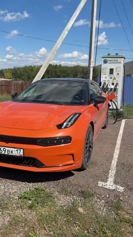 The video shows an orange car parked next to a charging station. The car is facing the camera and is in focus. The charging station is to the right of the car, and there is a pole behind the car. The background shows a blue sky with clouds, trees, and mountains. There are also some buildings in the distance. The car's license plate is visible and reads "0969 BK 17". The charging station has the logo of "РОССЕТИ" on it. The video is shot in daylight.