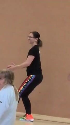 A woman with glasses and her hair in a ponytail, wearing a black t-shirt and black leggings with rainbow stripes, is dancing in front of a brown wall. She is performing a series of dance moves, including arm gestures and leg movements. There is a young child in a white uniform on the left side of the frame, and two other people partially visible on the right. The floor is light colored.