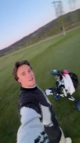 A young man films a selfie video on a golf course. The sky is a light blue and it appears to be early evening. There are mountains and trees visible in the background. The man is wearing a patterned sweater. He is walking and filming his golf bag on a three-wheeled push cart. There are other people playing golf in the distance.