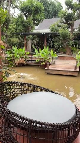 A video pans across a tranquil outdoor scene. In the foreground, a round table with a gray surface is nestled within a dark, woven wicker chair. Behind the chair, a murky pond reflects the overcast sky and lush greenery. A small fountain in the pond sprays water, creating ripples. Wooden decking and walkways surround the pond, leading to a building with a dark roof partially obscured by trees. Large green plants with broad leaves grow along the water's edge. The overall impression is one of a peaceful, natural setting, possibly a garden cafe or resort. No people are visible.