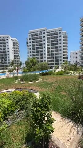 This is a landscape video shot on a sunny day, likely in the morning or early afternoon, given the bright sunlight and clear blue sky. The scene depicts a modern residential complex with several multi-story white buildings featuring numerous balconies. In the foreground, there is a landscaped garden with lush green grass, various bushes, ornamental plants, and some flowering plants in shades of pink and red. A pathway made of light-colored paving stones is visible to the right. In the middle ground, a swimming pool with blue water is visible, and a few people can be seen near it, suggesting leisure activities. The overall setting appears to be a resort or a contemporary apartment complex in a warm climate. The architecture of the buildings, the landscaping, and the clear, bright sky suggest a location in a Mediterranean or similar region, aligning with the provided location context of Trikomo, Northern Cyprus. There is no visible text in the video. The mood conveyed is one of relaxation and modern living, with the sunny weather enhancing the pleasant atmosphere.