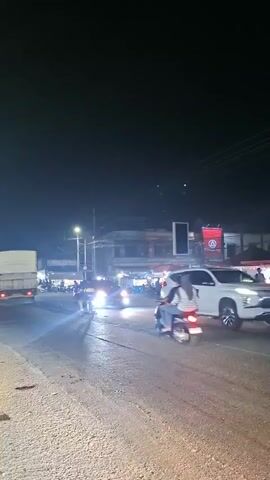 The video shows a nighttime street scene. In the foreground, the asphalt road is visible, wet from rain or moisture, with tire tracks. A dark-colored minivan is the central focus of the latter half of the video, with its taillights illuminated and blue light emanating from beneath it. The minivan has tinted windows, and a blue light is visible on its roof. In the background, there are several buildings with illuminated signs, suggesting a commercial or town area. Streetlights are on, casting light onto the road and surrounding buildings. Other vehicles, including a white SUV and motorcycles, are present on the road, along with a few pedestrians. The overall atmosphere is one of a busy street at night.