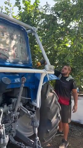 A man is seen in front of a blue tractor, holding a yellow and black tool, possibly a heat gun or a small blower, and aiming it at the side of the tractor. He is wearing a black and red t-shirt and black shorts, with a white cloth draped over his shoulder. The tractor's windows are covered with paper. The scene takes place outdoors, with trees and foliage visible in the background. The man appears to be working on the tractor, perhaps preparing it for painting or drying a substance.