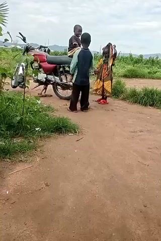 The video shows three children and a motorcycle on a dirt road. Two boys are standing near a red motorcycle, one of whom is seated on it. A girl in a colorful dress stands to the right of the motorcycle. The children appear to be interacting with each other. The background consists of green vegetation and a cloudy sky.