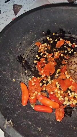 A close-up shot shows a hand holding a pestle and grinding red chilies in a dark mortar. The chilies are chopped and scattered around the mortar, with their seeds visible. The pestle is made of wood and has a textured surface. The mortar is made of dark stone. The background is blurry but appears to be a countertop with some other objects. The lighting is dim.