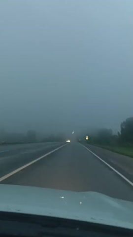 The video shows a first-person perspective of driving on a highway in dense fog. The road ahead is visible, with lane markings and a yellow line separating lanes. Other vehicles are visible in the distance, their headlights cutting through the fog. The environment is overcast and appears to be rural, with trees and vegetation visible on the sides of the road, obscured by the fog. The video captures the feeling of limited visibility and the caution required when driving in such conditions.