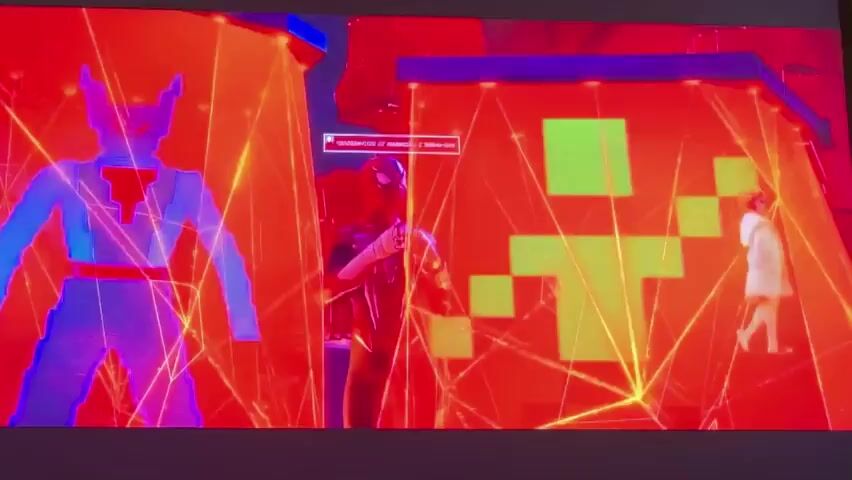 This media file appears to be a clip from an animated film, likely "Spider-Man: Into the Spider-Verse" given the visual style and character designs. The scene is vibrant and abstract, dominated by a striking red and orange color palette with geometric, crystalline structures overlaying the visuals, suggesting a surreal or dimensional setting.

The primary subjects are animated characters who appear to be in a tense or significant moment. In the foreground left, a young Black woman with elaborate, gravity-defying hair is seen in profile, wearing a stylish outfit with a star motif. To her right, a young Black man, also in profile, is looking towards the viewer with a concerned expression. He is wearing a black suit with a red spider emblem. Another young woman with blonde hair stands next to him. In the background, other figures are visible, some silhouetted, including a light-skinned figure in a white outfit standing on a platform and a robotic-looking character.

The activity suggested is one of confrontation or transition. The characters seem to be reacting to something happening around them, and the overall atmosphere is charged with emotion, possibly anxiety or determination. The fragmented and glowing nature of the environment could represent a breakdown of reality, a portal, or a digital space.

Visible text appears on screen in Russian, overlaid on the scene. The text reads "Шёл бы ты курсивом" which translates to "You would go in italics". Below this, it reads "человек-паук!" which translates to "Spider-Man!". This text likely refers to the character and perhaps a playful or taunting statement directed at him. The media seems to be presented on a large screen, possibly in a public viewing or as part of a demonstration. There are no discernible cues for time of day or weather as the setting is entirely abstract and artificial.