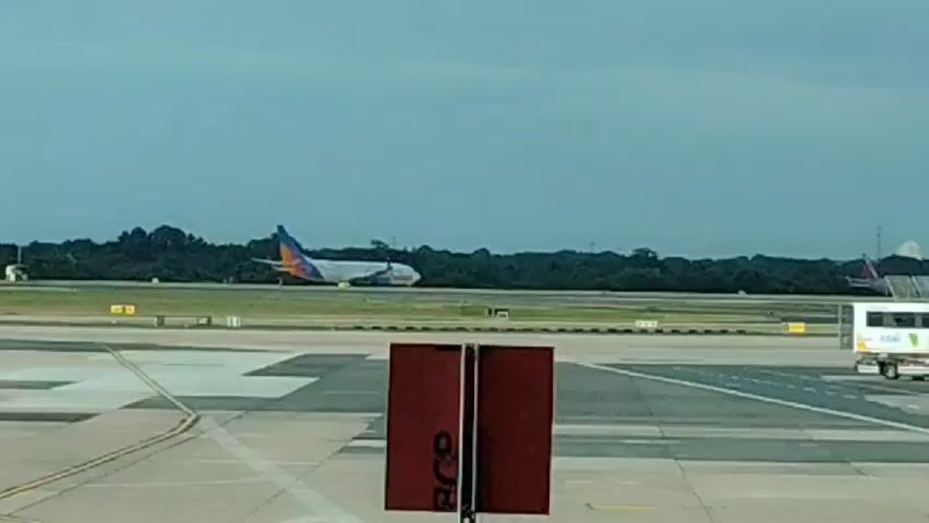 The video shows an airport tarmac under a cloudy sky. In the background, a passenger airplane with a white fuselage and a blue and orange tail is visible on a runway, positioned at an angle. The plane appears to be stationary. To the right of the plane, there is a white vehicle with green accents, possibly a baggage tug or shuttle bus, also on the tarmac. In the foreground, two red vertical posts with white lettering are positioned in the center of the frame, framing the runway and the background. The tarmac is marked with yellow and white lines, indicating taxiways and runways. Dense green trees form a backdrop behind the runway, and a faint structure resembling a distant control tower or building is visible on the far right horizon. The overall impression is a static scene at an airport, likely during a period of aircraft movement or waiting.