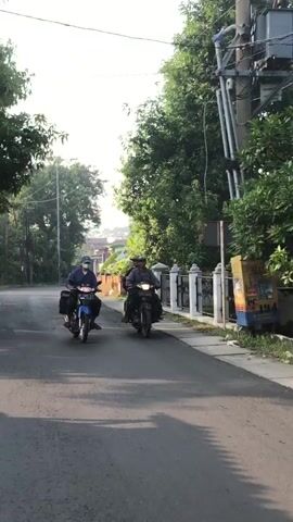 Two people are riding motorcycles down a paved road lined with trees and a white fence. The person on the left is wearing a blue shirt and a hat, while the person on the right is wearing a dark shirt and a hat. Both are wearing masks. The motorcycle on the left is blue and the motorcycle on the right is black. There is a utility pole with wires on the right side of the road. The sun is shining and casting shadows on the road.