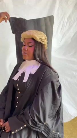A young woman dressed in barrister attire sits in a chair, posing for a photograph. She is wearing a black gown, a white jabot, and a traditional blonde barrister's wig over her long dark hair. A dark-skinned hand is visible in the upper left of the frame, holding a black fabric piece. The background consists of a white draped sheet with some black objects taped to it. A man in a red and black plaid shirt walks past the woman on her right side.