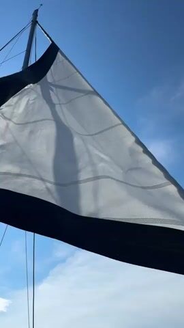A close-up shot of a white sail with dark blue trim on a sailboat, captured against a bright blue sky with scattered clouds. The sail is billowed by the wind and attached to a mast. The sun's rays create shadows on the sail, and rigging lines are visible. The camera angle looks upwards, emphasizing the sail against the sky.