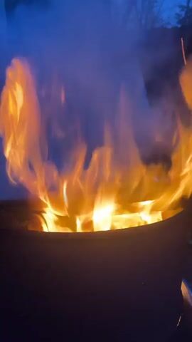 A close-up shot of a vigorous fire burning within a dark, circular fire pit or container. Bright orange and yellow flames rise high, producing a significant amount of smoke that billows upwards and obscures parts of the background. The flames flicker and dance intensely. In the blurred background, silhouetted tree branches and a dark blue sky suggest it is either dusk or nighttime. No people or text are visible in the video. The scene takes place outdoors, possibly in Ильинская Поляна Снт, Russia, as indicated by the location context.